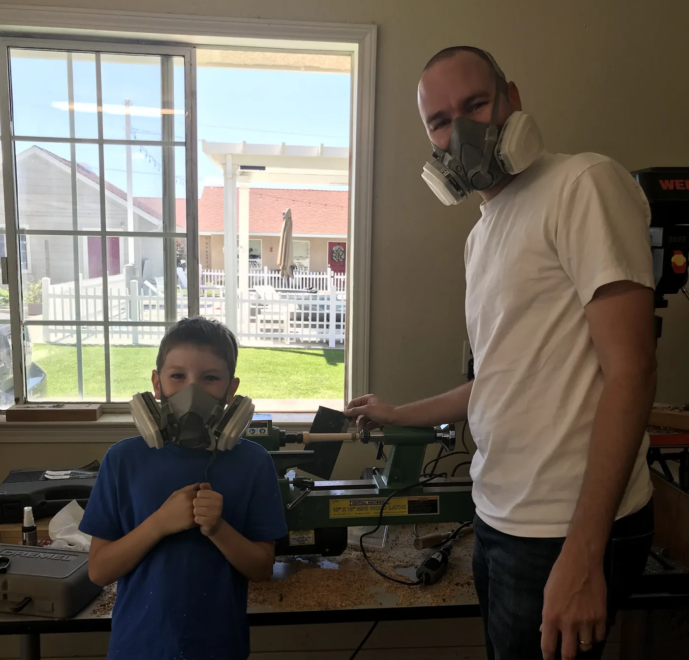 A father and his son in respirators standing together beside the wood lathe in their home shop